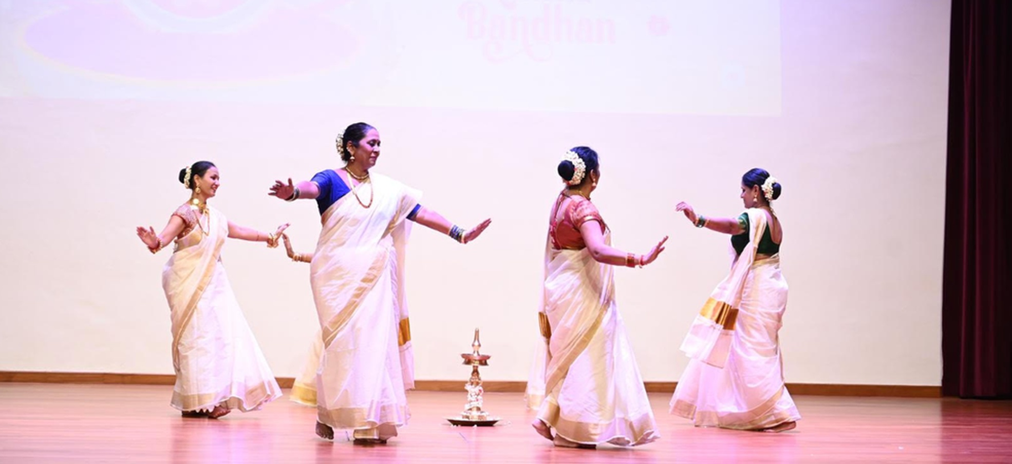 The Taranga dance troupe performing a Thiruvathira-style Kerala dance in white sarees with gold borders, around the traditional brass lamp