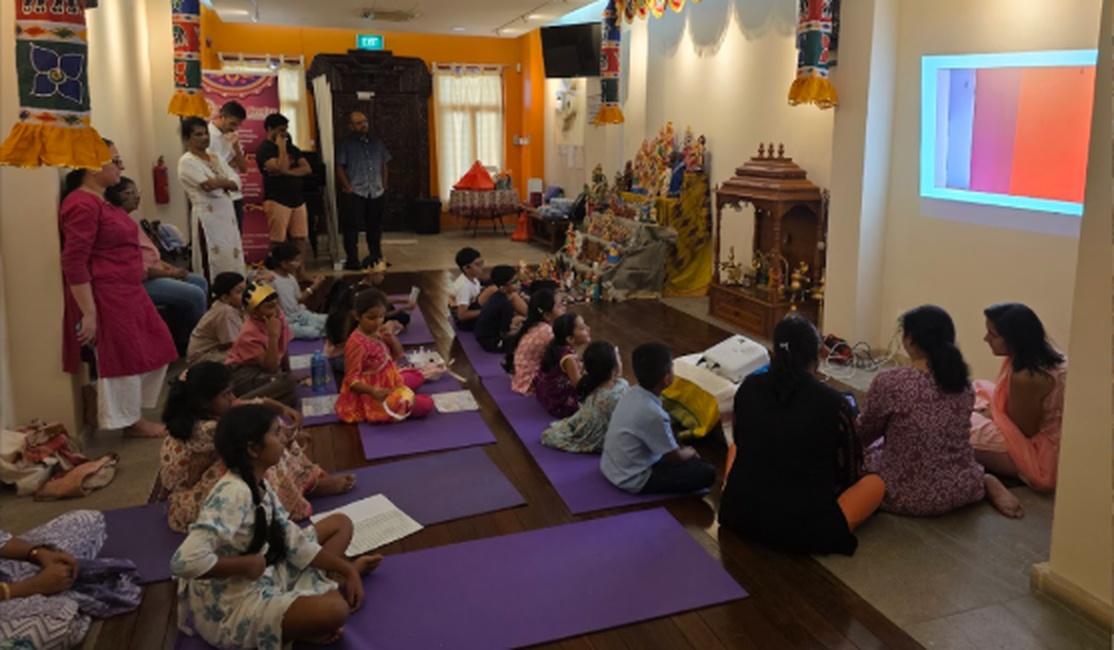 Balagurukulam children seated on purple yoga mats in the Hindu Centre hall, with the deity altar at the back and decorative banners overhead