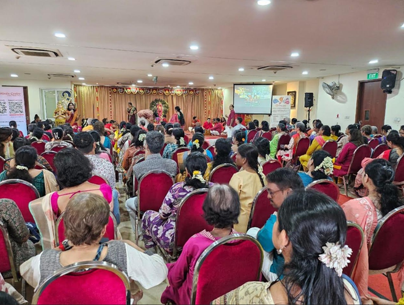 A packed auditorium audience at a Hindu Centre lecture event, with stage performance and screen visible at the front