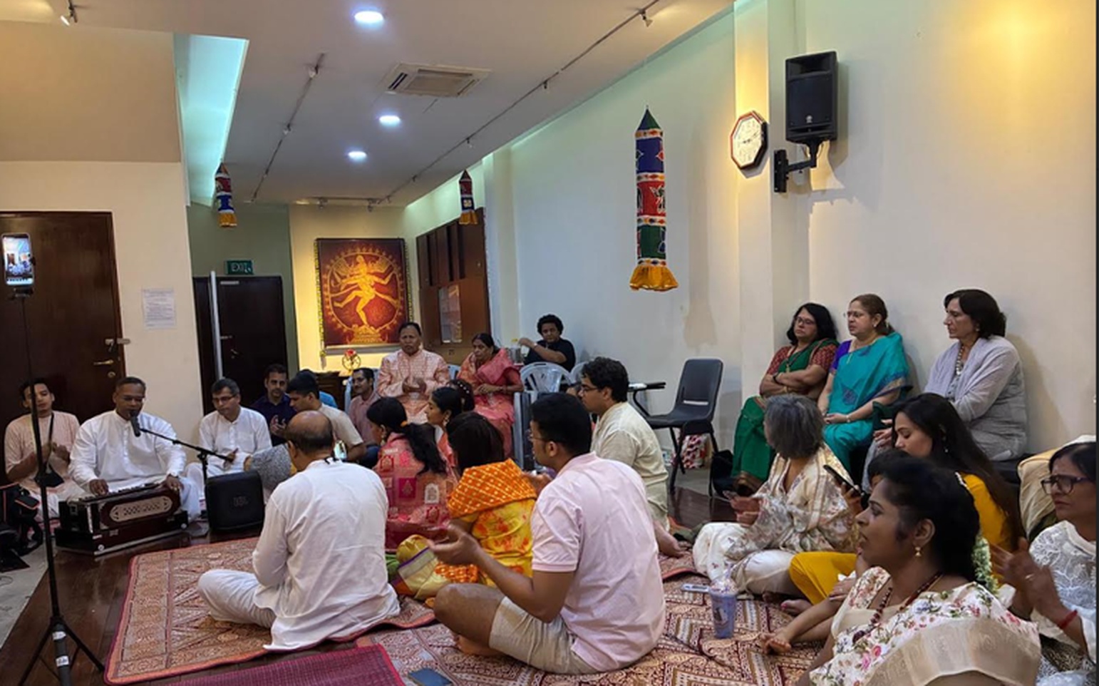 Devotees seated for a chanting and bhajan session at Hindu Centre, harmonium player at front, with Nataraja image behind